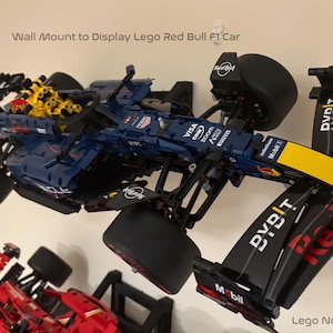May include: Wall mount for displaying a Lego Red Bull Formula One race car. The car is blue, black, and yellow with the Red Bull logo and sponsors' logos. The mount is black and has a clear plastic backing.