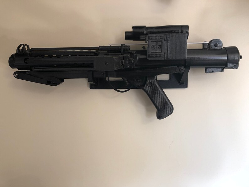 May include: A black plastic toy blaster with a scope and a handle. The blaster is mounted on a wall.