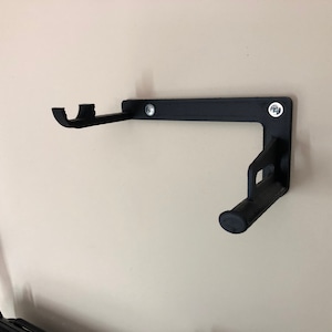 May include: A black, wall-mounted bracket with two curved supports and a cylindrical end. The bracket is secured to the wall with two screws. The design is simple and functional, likely for holding an item.