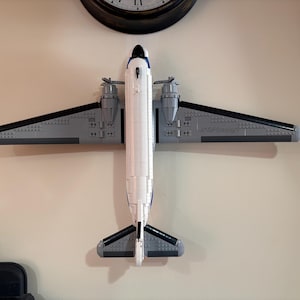 May include: A model airplane made of gray, white, and black blocks is mounted on a beige wall. The airplane has two propellers, wings, and a tail. The text "JASFDesign" is visible on the wing. A black clock is above the airplane.