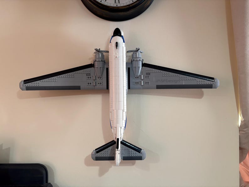 May include: A model airplane made of gray, white, and black blocks is mounted on a beige wall. The airplane has two propellers, wings, and a tail. The text "JASFDesign" is visible on the wing. A black clock is above the airplane.