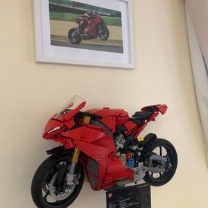 May include: A red Lego Ducati Panigale V4 R motorcycle model mounted on a wall. The model is detailed with black wheels, a black exhaust pipe, and a black seat. The model is displayed with a black plaque with the Ducati logo and specifications.