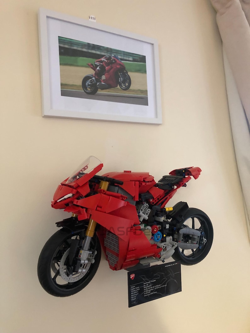 May include: A red Lego Ducati Panigale V4 R motorcycle model mounted on a wall. The model is detailed with black wheels, a black exhaust pipe, and a black seat. The model is displayed with a black plaque with the Ducati logo and specifications.