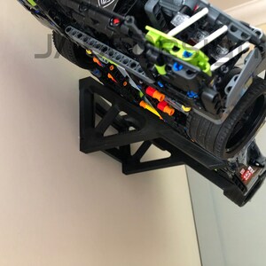 Pu&ograve; includere: Un modello di auto Lego nero montato su un supporto da parete nero. L'auto &egrave; capovolta e ha uno schema di colori verde e nero. Il supporto da parete ha una forma triangolare.