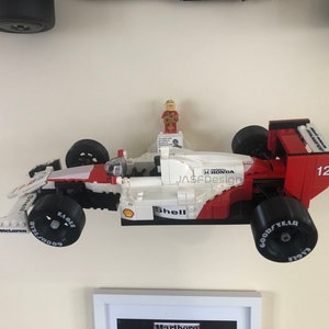 Op de afbeelding: Een Lego-model van een Formule 1-raceauto, voornamelijk wit en rood, met zwarte banden. De auto heeft sponsorlogo's zoals Shell, Goodyear en McLaren. Een klein Lego-figuur staat bovenop. Het nummer 12 is zichtbaar.