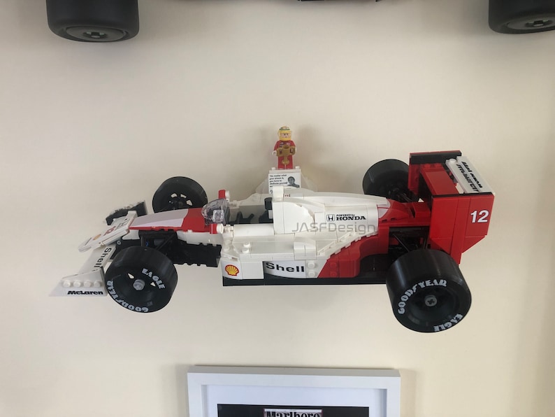 Op de afbeelding: Een Lego-model van een Formule 1-raceauto, voornamelijk wit en rood, met zwarte banden. De auto heeft sponsorlogo's zoals Shell, Goodyear en McLaren. Een klein Lego-figuur staat bovenop. Het nummer 12 is zichtbaar.