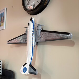 May include: A model airplane made of gray, white, and black blocks, mounted on a wall. The plane has the words "PAN AMERICAN" and a logo on the tail. The wings are gray with black edges. A clock is visible in the background.