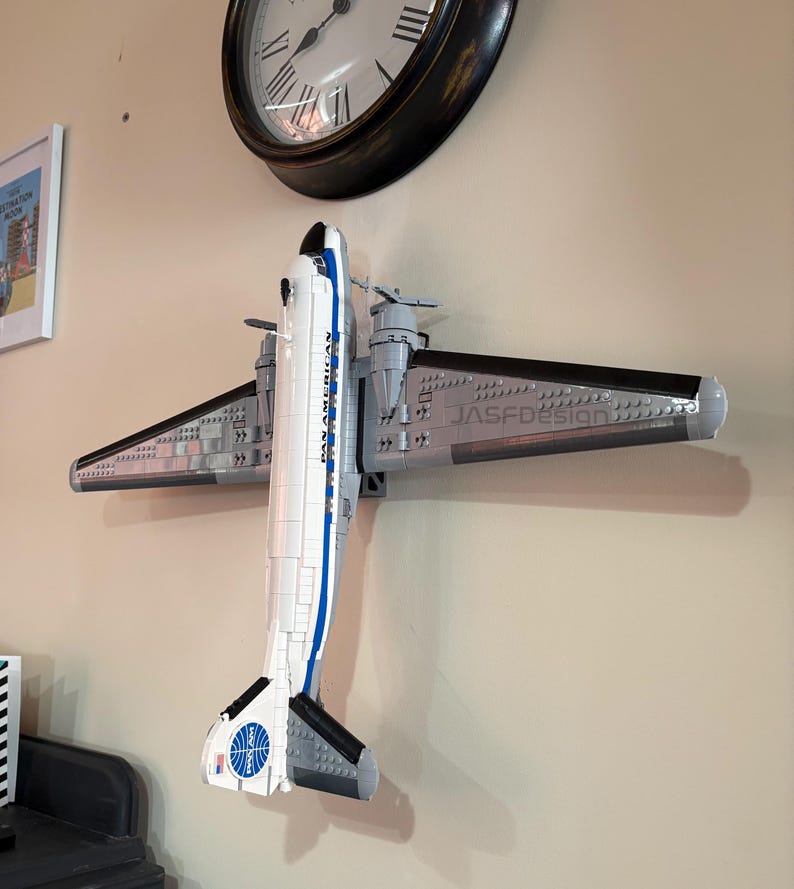 May include: A model airplane made of gray, white, and black blocks, mounted on a wall. The plane has the words "PAN AMERICAN" and a logo on the tail. The wings are gray with black edges. A clock is visible in the background.