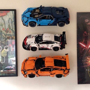 May include: Three Lego car models displayed on a white wall. The cars are a blue Bugatti Chiron, a white Porsche 911 GT3 RS, and an orange Porsche 911 GT3 RS.