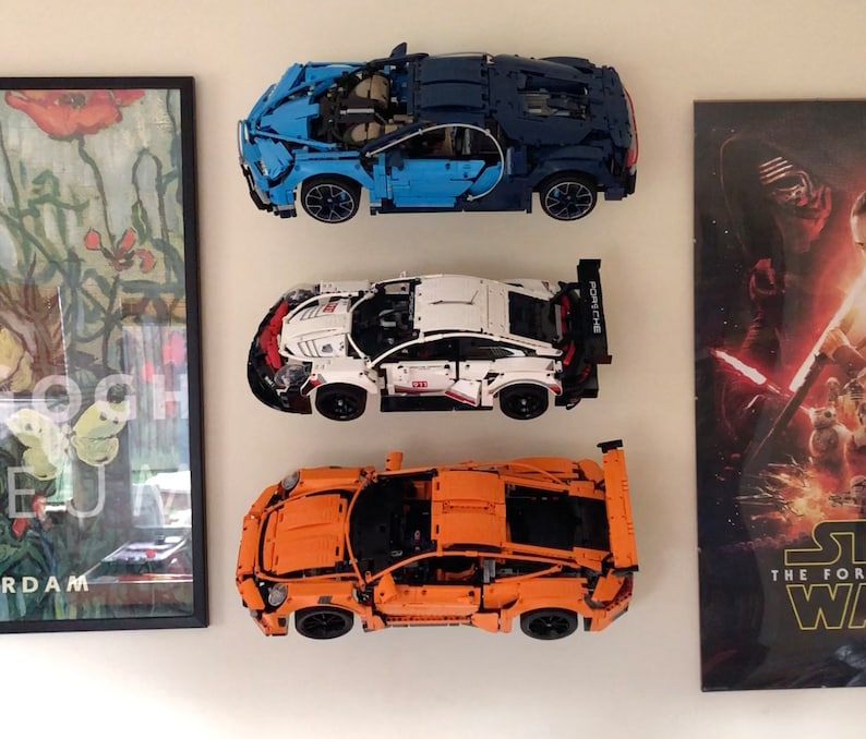 May include: Three Lego car models displayed on a white wall. The cars are a blue Bugatti Chiron, a white Porsche 911 GT3 RS, and an orange Porsche 911 GT3 RS.