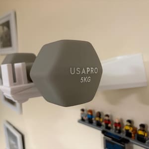 Puede incluir: Una pesa gris de 5 kg, marcada "USAPRO", est&aacute; montada en un soporte blanco. La pesa tiene forma hexagonal. El soporte est&aacute; montado en una pared beige. Debajo hay una peque&ntilde;a estanter&iacute;a con figuritas.