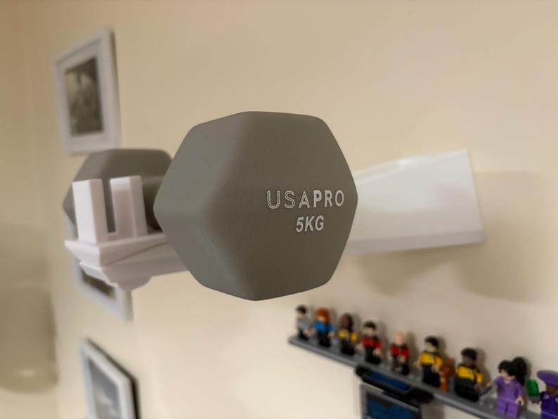 Puede incluir: Una pesa gris de 5 kg, marcada "USAPRO", est&aacute; montada en un soporte blanco. La pesa tiene forma hexagonal. El soporte est&aacute; montado en una pared beige. Debajo hay una peque&ntilde;a estanter&iacute;a con figuritas.