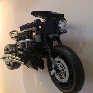 May include: A black Lego motorcycle model with a clear headlight and a detailed engine. The motorcycle is mounted on a white wall.