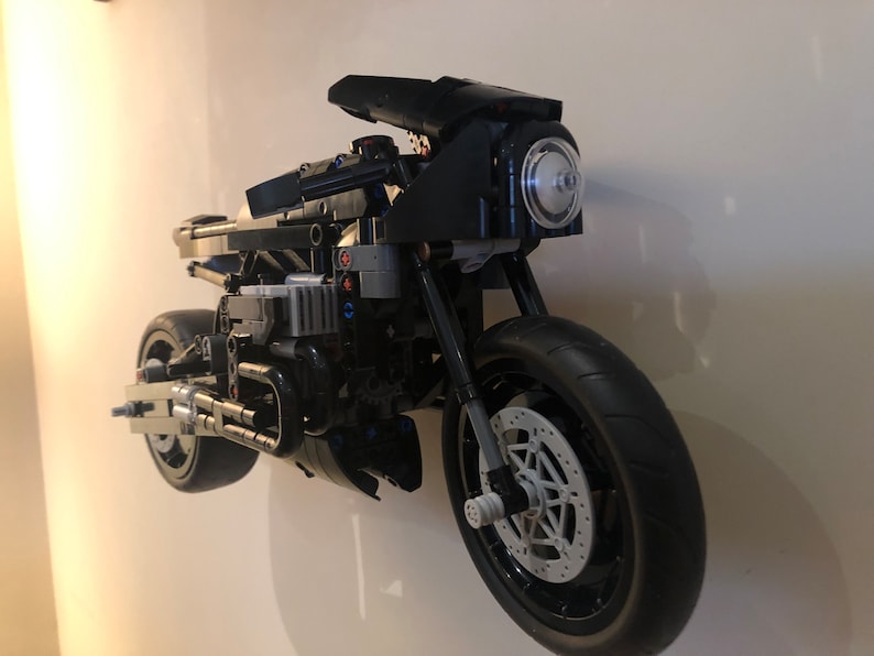 May include: A black Lego motorcycle model with a clear headlight and a detailed engine. The motorcycle is mounted on a white wall.