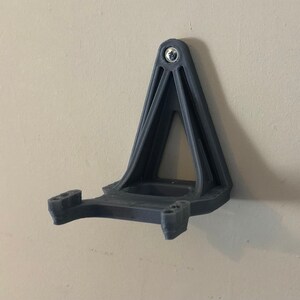 May include: A 3D printed wall mount for a VR headset, made of black plastic. The mount has a triangular base and a curved arm to hold the headset.