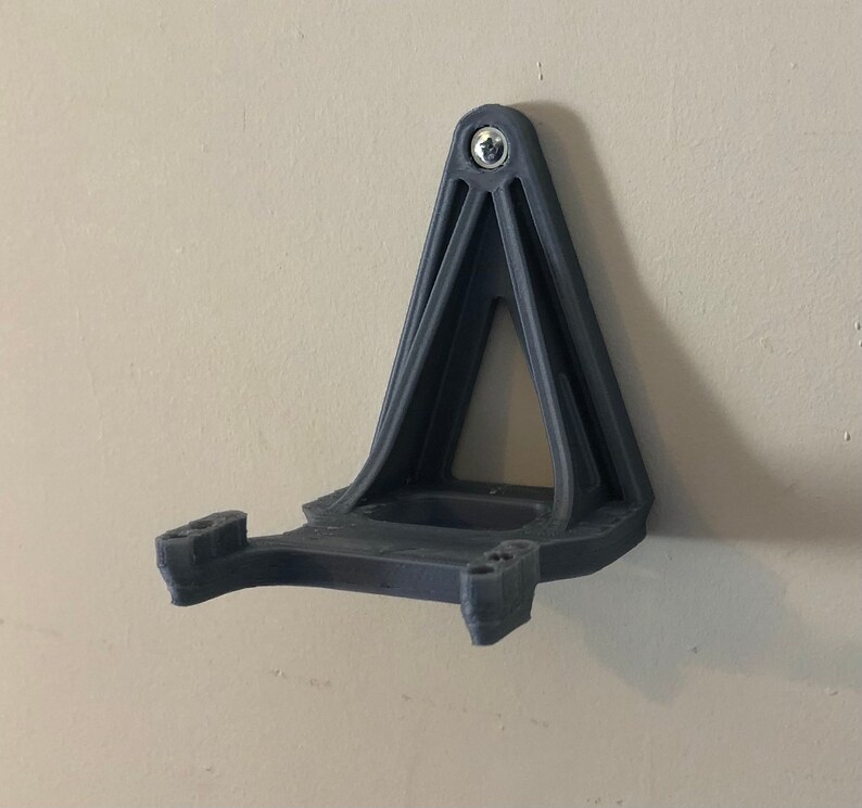 May include: A 3D printed wall mount for a VR headset, made of black plastic. The mount has a triangular base and a curved arm to hold the headset.
