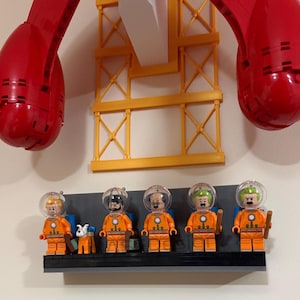 Puede incluir: Una exhibici&oacute;n de figuras de astronautas de Lego con trajes naranjas y cascos transparentes, de pie sobre una estanter&iacute;a negra. Tambi&eacute;n hay una peque&ntilde;a figura de perro. El fondo incluye una estructura de gr&uacute;a amarilla y grandes piezas rojas de Lego.