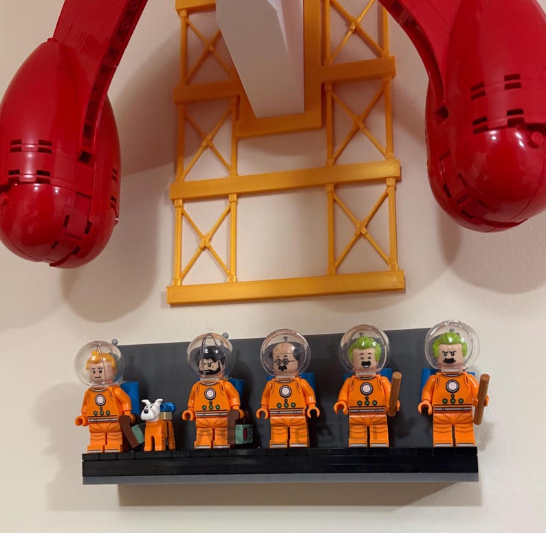 Puede incluir: Una exhibici&oacute;n de figuras de astronautas de Lego con trajes naranjas y cascos transparentes, de pie sobre una estanter&iacute;a negra. Tambi&eacute;n hay una peque&ntilde;a figura de perro. El fondo incluye una estructura de gr&uacute;a amarilla y grandes piezas rojas de Lego.