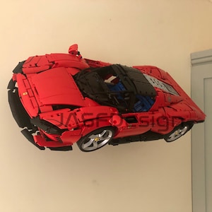 May include: A red Lego model of a Ferrari sports car with black accents and a black roof. The car is suspended in mid-air against a white wall.