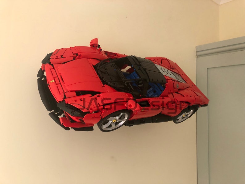 May include: A red Lego model of a Ferrari sports car with black accents and a black roof. The car is suspended in mid-air against a white wall.