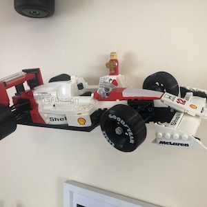 Op de afbeelding: Een wit, rood en zwart Lego-model van een Formule 1-raceauto, aan een muur bevestigd. De auto heeft nummer 12, Shell- en Goodyear-merken en een klein Lego-figuur. De tekst "JASFDesign" is zichtbaar.
