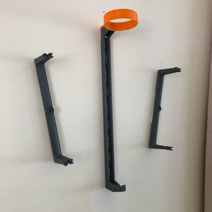 May include: Three wall-mounted black plastic holders for a 3D printer filament spool. The top holder has an orange ring.