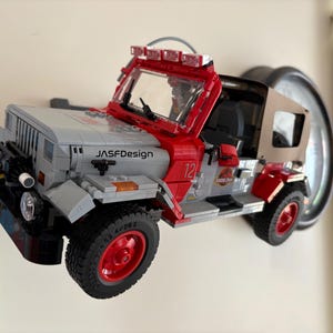 May include: A gray and red toy off-road vehicle model featuring a tan soft top, a front windshield with a light bar, and the text "JASFDesign" on the hood. The vehicle has red wheels, black tires, and a Jurassic Park logo on the side door panel.
