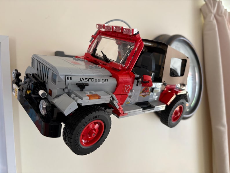May include: A gray and red toy off-road vehicle model featuring a tan soft top, a front windshield with a light bar, and the text "JASFDesign" on the hood. The vehicle has red wheels, black tires, and a Jurassic Park logo on the side door panel.