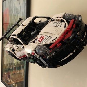 May include: A white and red Lego Technic Porsche 911 RSR race car model with black accents and a Mobil 1 logo. The car is mounted on a wall and has a rear wing.