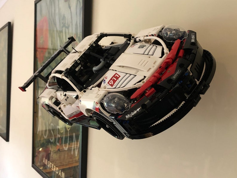 May include: A white and red Lego Technic Porsche 911 RSR race car model with black accents and a Mobil 1 logo. The car is mounted on a wall and has a rear wing.