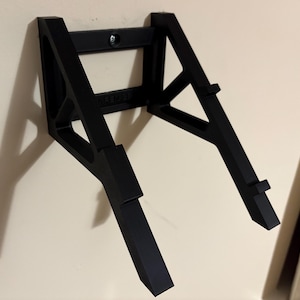 May include: A black wall-mounted guitar stand. The stand is made of a sturdy material and features a rectangular top section with a mounting screw and two angled support arms. The arms have small ledges to hold the guitar.