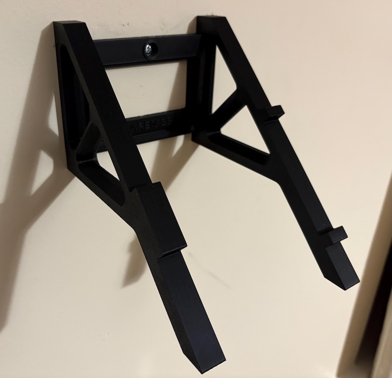 May include: A black wall-mounted guitar stand. The stand is made of a sturdy material and features a rectangular top section with a mounting screw and two angled support arms. The arms have small ledges to hold the guitar.