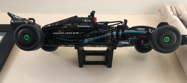 May include: A black and silver Lego model of a Formula 1 race car with the Petronas logo and Ineos branding. The car is mounted on a black stand.