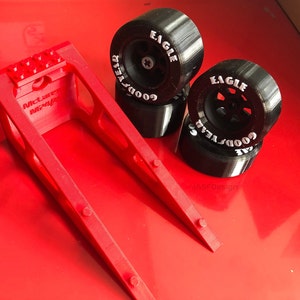 Op de afbeelding: Een rode 3D-geprinte standaard met de tekst "McLaren MP4/4" en vier zwarte Goodyear Eagle racebanden. De banden hebben witte letters en zijn gestapeld op een rode ondergrond.