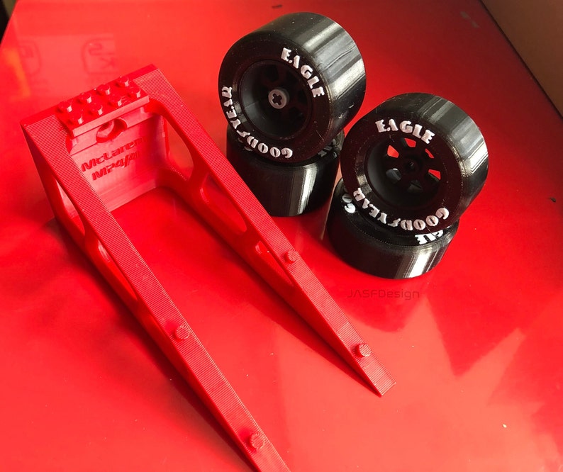 Op de afbeelding: Een rode 3D-geprinte standaard met de tekst "McLaren MP4/4" en vier zwarte Goodyear Eagle racebanden. De banden hebben witte letters en zijn gestapeld op een rode ondergrond.