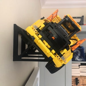 K&ouml;nnte beinhalten: Ein schwarzer 3D-gedruckter Wandhalter, der f&uuml;r ein gelbes Lego-Baufahrzeug ausgelegt ist. Das Fahrzeug ist kopf&uuml;ber an der Wandhalterung montiert.