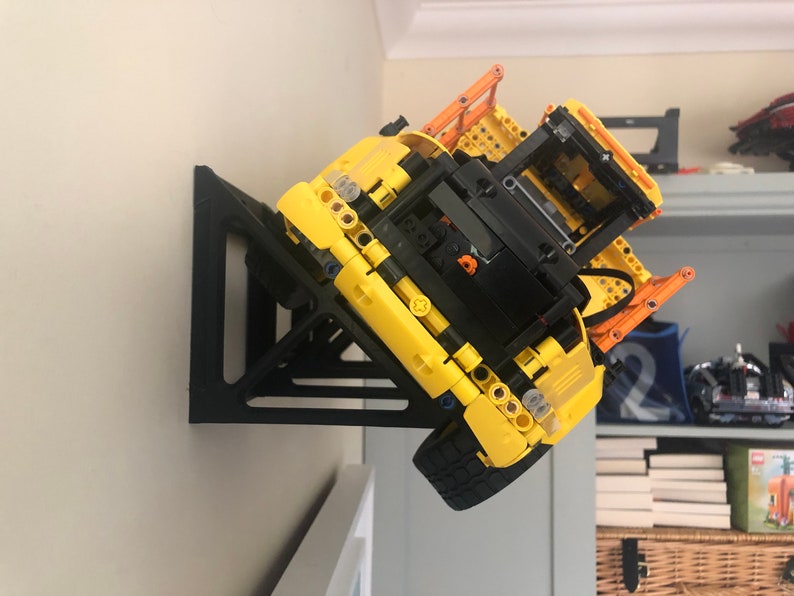 K&ouml;nnte beinhalten: Ein schwarzer 3D-gedruckter Wandhalter, der f&uuml;r ein gelbes Lego-Baufahrzeug ausgelegt ist. Das Fahrzeug ist kopf&uuml;ber an der Wandhalterung montiert.