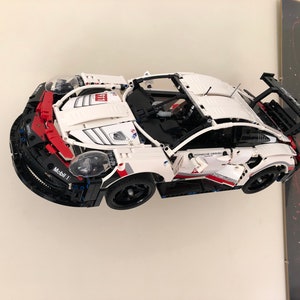May include: A white and black Lego Porsche 911 RSR race car with red accents and a Mobil 1 logo. The car is mounted on a wall with a Star Wars poster in the background.