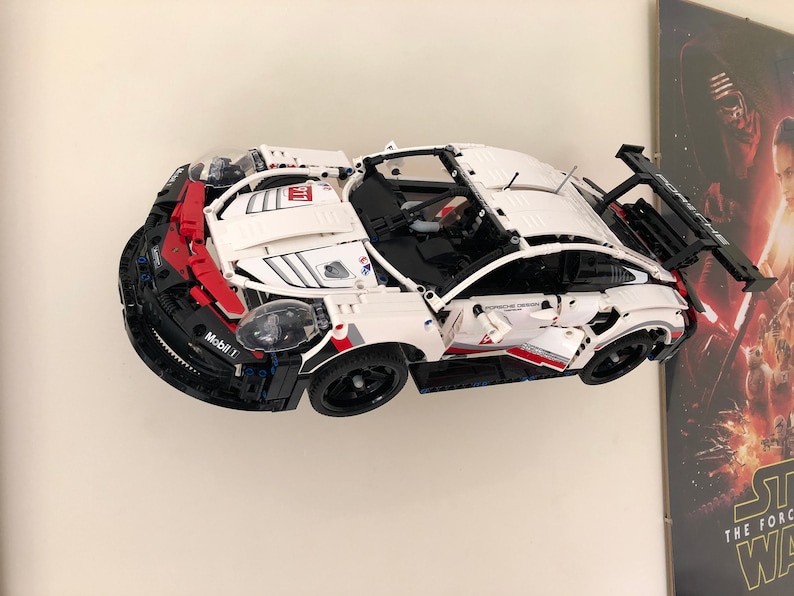 May include: A white and black Lego Porsche 911 RSR race car with red accents and a Mobil 1 logo. The car is mounted on a wall with a Star Wars poster in the background.