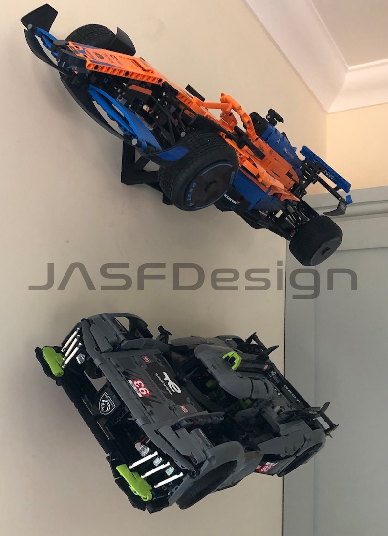 May include: Two Lego race cars mounted on a wall. The orange and blue car is a Formula 1 race car. The black and green car is a sports car. The cars are displayed on a white wall.