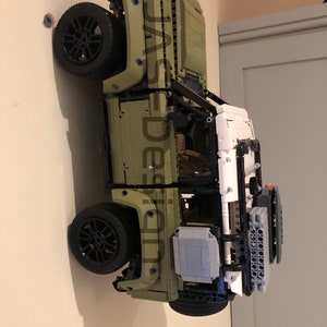 May include: A green Lego Land Rover Defender model with black wheels and a white roof. The model is mounted on a wall.