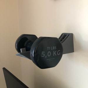 May include: Black dumbbell weight rack mounted on a wall with a 11 lbs / 5.0 kg dumbbell in place.