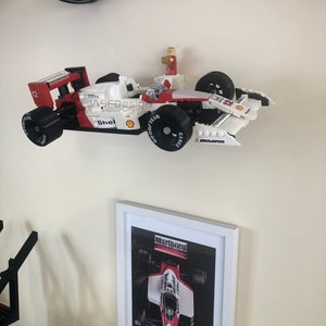 Op de afbeelding: Een wit en rood Lego raceauto model met zwarte banden en het nummer 12. Het model heeft Shell- en Goodyear-logo's. Een ingelijste foto van een raceauto staat eronder. De tekst "Marlboro" is zichtbaar.