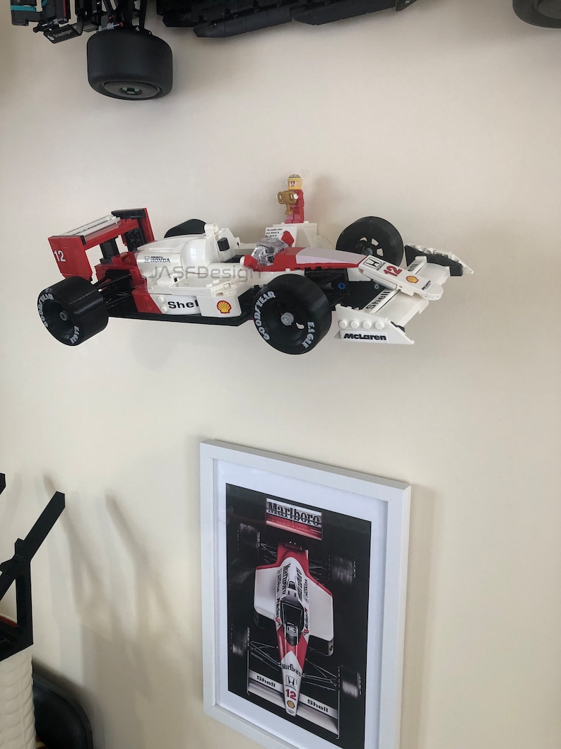 Op de afbeelding: Een wit en rood Lego raceauto model met zwarte banden en het nummer 12. Het model heeft Shell- en Goodyear-logo's. Een ingelijste foto van een raceauto staat eronder. De tekst "Marlboro" is zichtbaar.