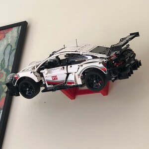 May include: A white, black, and red Lego Porsche 911 GT3 RS race car model is mounted on a red wall mount. The car is suspended in mid-air, as if it is racing.