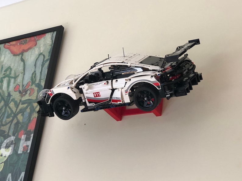 May include: A white, black, and red Lego Porsche 911 GT3 RS race car model is mounted on a red wall mount. The car is suspended in mid-air, as if it is racing.