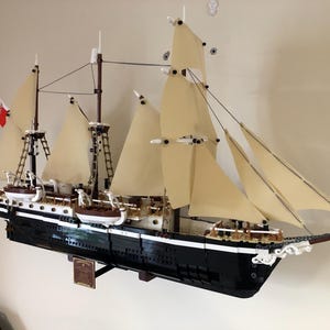 May include: A detailed model of a black and white sailing ship with white sails, built from LEGO bricks. The ship is mounted on a wooden stand and is displayed on a wall.