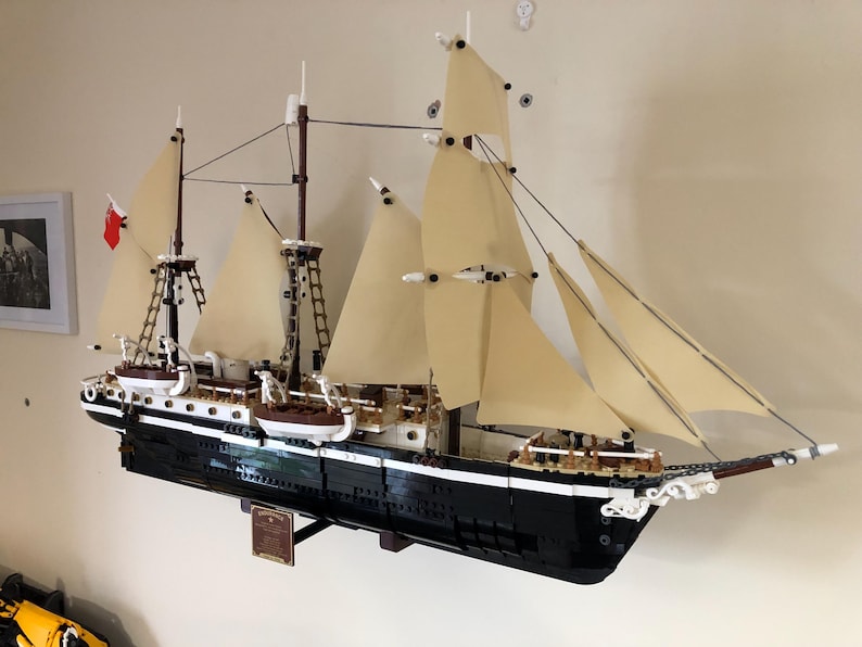 May include: A detailed model of a black and white sailing ship with white sails, built from LEGO bricks. The ship is mounted on a wooden stand and is displayed on a wall.
