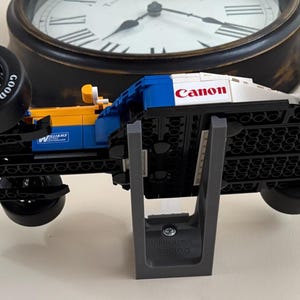 May include: A Lego race car model in blue, yellow, and white, with "Canon" printed on the top. The car has black tires with "Goodyear" and "Eagle" branding. The model is displayed on a gray stand.