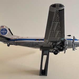 May include: A model airplane in the style of a vintage Pan Am airliner. The plane is primarily white and gray, with blue accents and the Pan Am logo on the tail. The wings are large and detailed, and the plane is mounted on a gray stand.